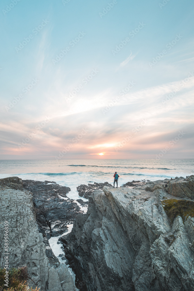 Fototapeta premium Fearless man with a backpack watching the sunset over the Atlantic Ocean in Porto Covo, Portugal. A hiker stands on the edge of a cliff looking into his future. Self-awareness