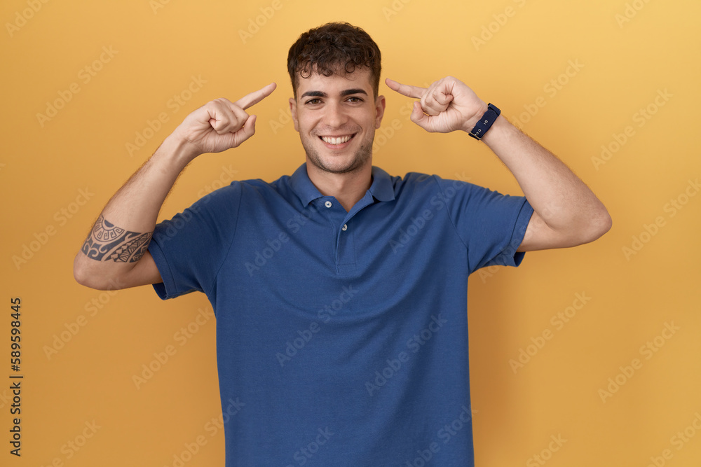 Young hispanic man standing over yellow background smiling pointing to head with both hands finger, great idea or thought, good memory Young hispanic man standing over yellow background smiling pointing to head with both hands finger, great idea or thought, good memory