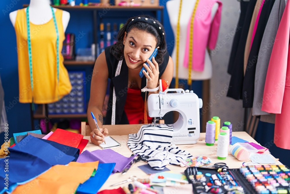 Fototapeta premium Young beautiful hispanic woman tailor talking on smartphone writing on notebook at clothing factory