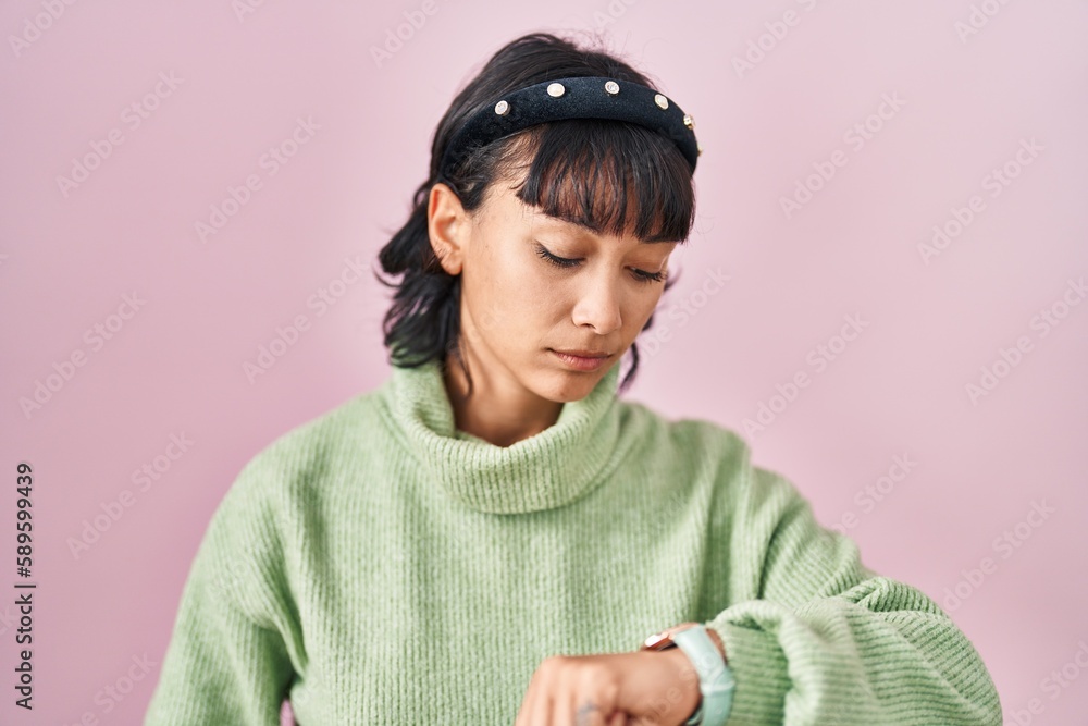 Young beautiful woman standing over pink background checking the time on wrist watch, relaxed and confident Young beautiful woman standing over pink background checking the time on wrist watch, relaxed and confident