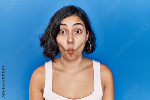 Young hispanic woman standing over blue background making fish face with lips, crazy and comical gesture. funny expression.