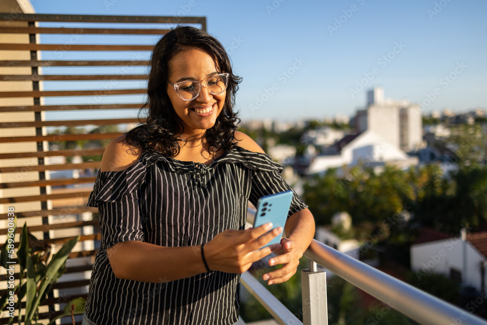 Portrait of smiling Latin woman with glasses using mobile phone on the balcony with view of the city