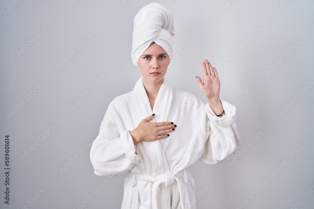 Blonde caucasian woman wearing bathrobe swearing with hand on chest and open palm, making a loyalty promise oath Blonde caucasian woman wearing bathrobe swearing with hand on chest and open palm, making a loyalty promise oath