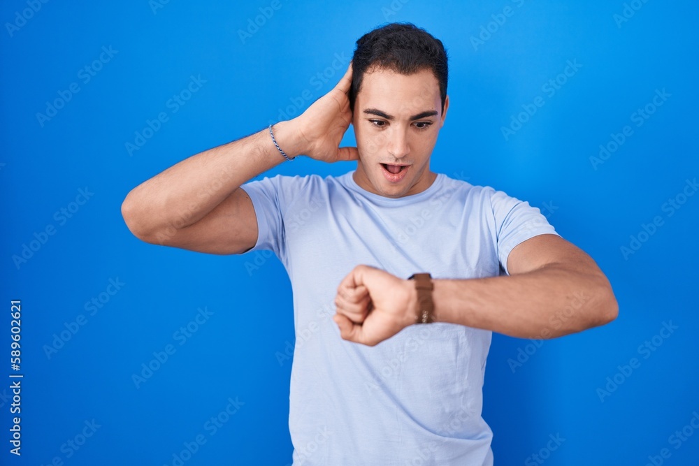 Young hispanic man standing over blue background looking at the watch time worried, afraid of getting late Young hispanic man standing over blue background looking at the watch time worried, afraid of getting late