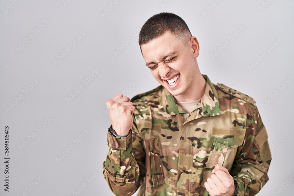 Young man wearing camouflage army uniform very happy and excited doing winner gesture with arms raised, smiling and screaming for success. celebration concept. Young man wearing camouflage army uniform very happy and excited doing winner gesture with arms raised, smiling and screaming for success. celebration concept.