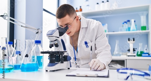 Young hispanic man scientist using microscope writing report at laboratory