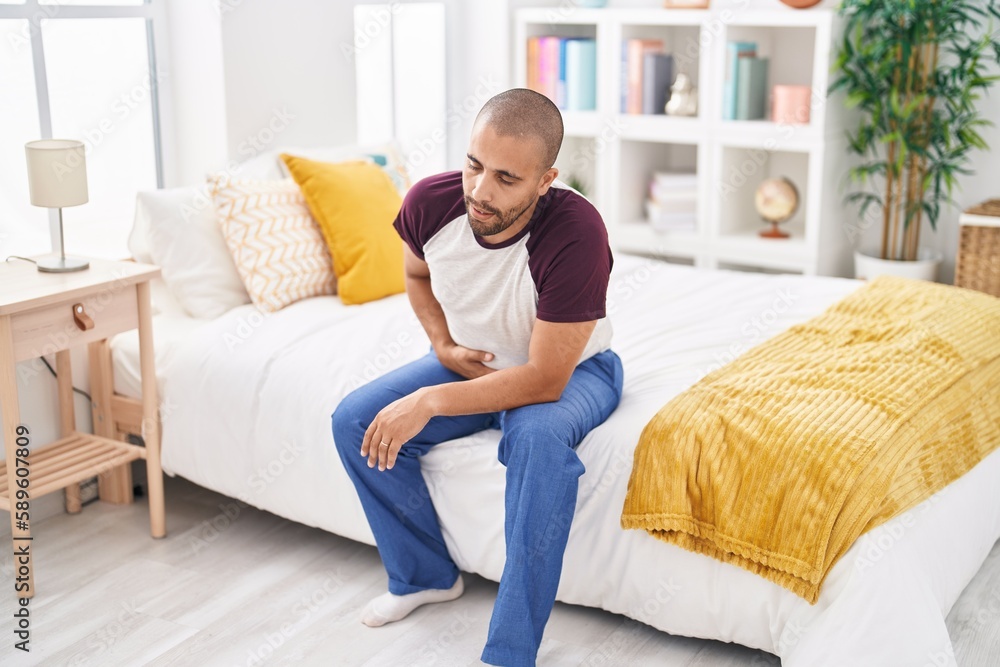 Young latin man suffering for stomach ache sitting on bed at bedroom Young latin man suffering for stomach ache sitting on bed at bedroom