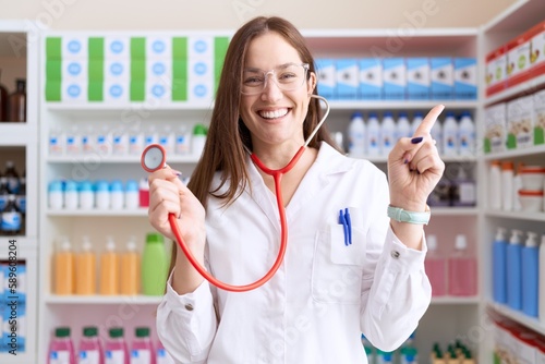 Beautiful brunette woman working at pharmacy drugstore holding stethoscope smiling happy pointing with hand and finger to the side