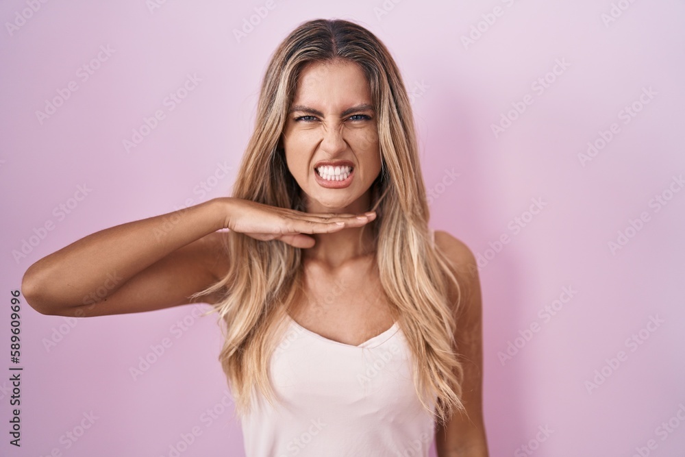 Young blonde woman standing over pink background cutting throat with hand as knife, threaten aggression with furious violence Young blonde woman standing over pink background cutting throat with hand as knife, threaten aggression with furious violence
