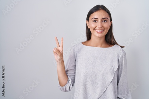 Young hispanic woman standing over white background showing and pointing up with fingers number two while smiling confident and happy.