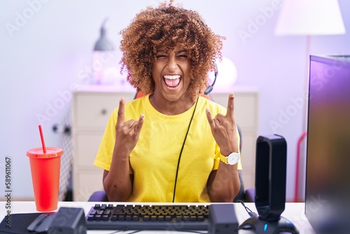 Young hispanic woman with curly hair playing video games wearing headphones shouting with crazy expression doing rock symbol with hands up. music star. heavy concept.