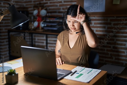 Young hispanic woman working at the office at night making fun of people with fingers on forehead doing loser gesture mocking and insulting.