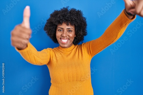 Black woman with curly hair standing over blue background approving doing positive gesture with hand, thumbs up smiling and happy for success. winner gesture.