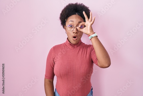 Beautiful african woman with curly hair standing over pink background doing ok gesture shocked with surprised face, eye looking through fingers. unbelieving expression.