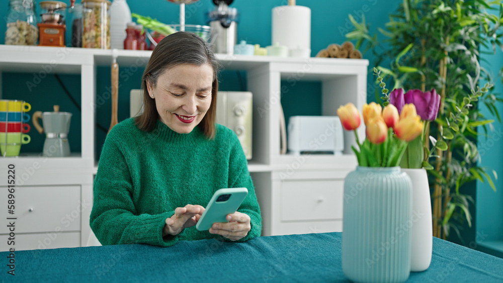 Mature hispanic woman with grey hair using smartphone sitting on the table at dinning room Mature hispanic woman with grey hair using smartphone sitting on the table at dinning room