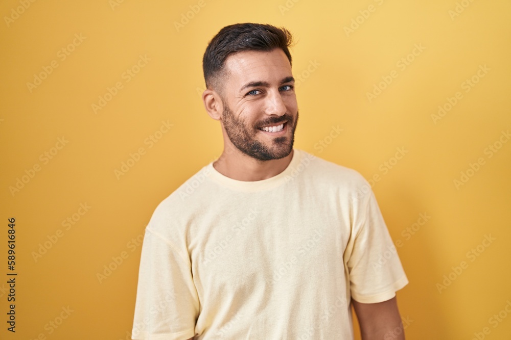 Handsome hispanic man standing over yellow background looking away to side with smile on face, natural expression. laughing confident. Handsome hispanic man standing over yellow background looking away to side with smile on face, natural expression. laughing confident.