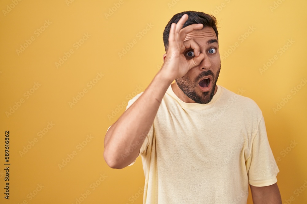 Handsome hispanic man standing over yellow background doing ok gesture shocked with surprised face, eye looking through fingers. unbelieving expression. Handsome hispanic man standing over yellow background doing ok gesture shocked with surprised face, eye looking through fingers. unbelieving expression.
