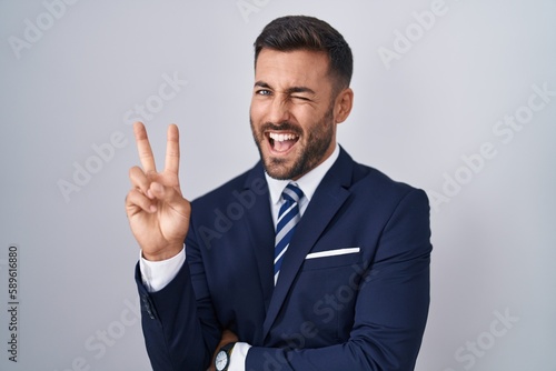Fotografie Handsome hispanic man wearing suit and tie smiling with happy face winking at the camera doing victory sign with fingers