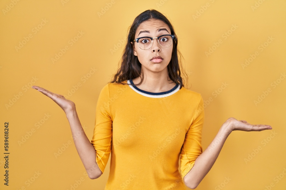 Young brazilian woman wearing glasses over yellow background clueless and confused expression with arms and hands raised. doubt concept. Young brazilian woman wearing glasses over yellow background clueless and confused expression with arms and hands raised. doubt concept.