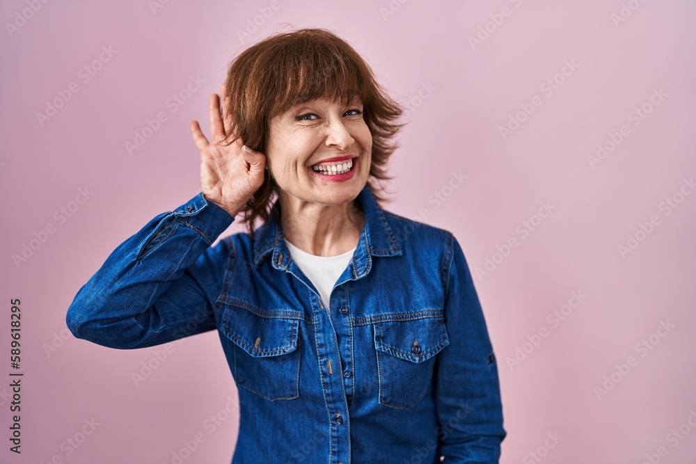 Middle age woman standing over pink background smiling with hand over ear listening an hearing to rumor or gossip. deafness concept. Middle age woman standing over pink background smiling with hand over ear listening an hearing to rumor or gossip. deafness concept.