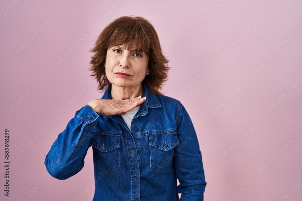 Middle age woman standing over pink background cutting throat with hand as knife, threaten aggression with furious violence Middle age woman standing over pink background cutting throat with hand as knife, threaten aggression with furious violence