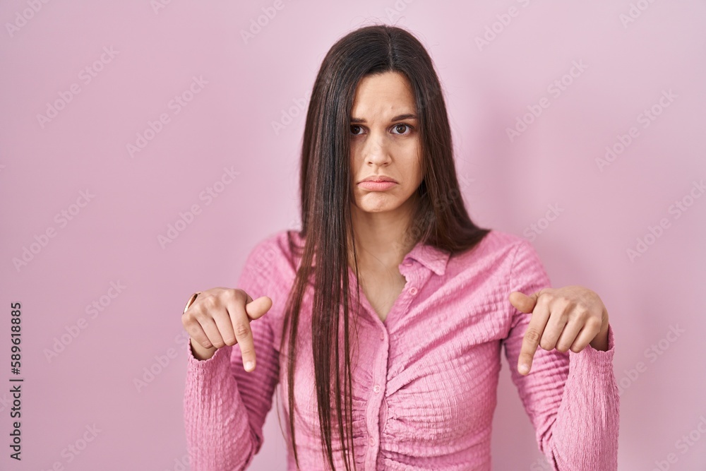Young hispanic woman standing over pink background pointing down looking sad and upset, indicating direction with fingers, unhappy and depressed. Young hispanic woman standing over pink background pointing down looking sad and upset, indicating direction with fingers, unhappy and depressed.