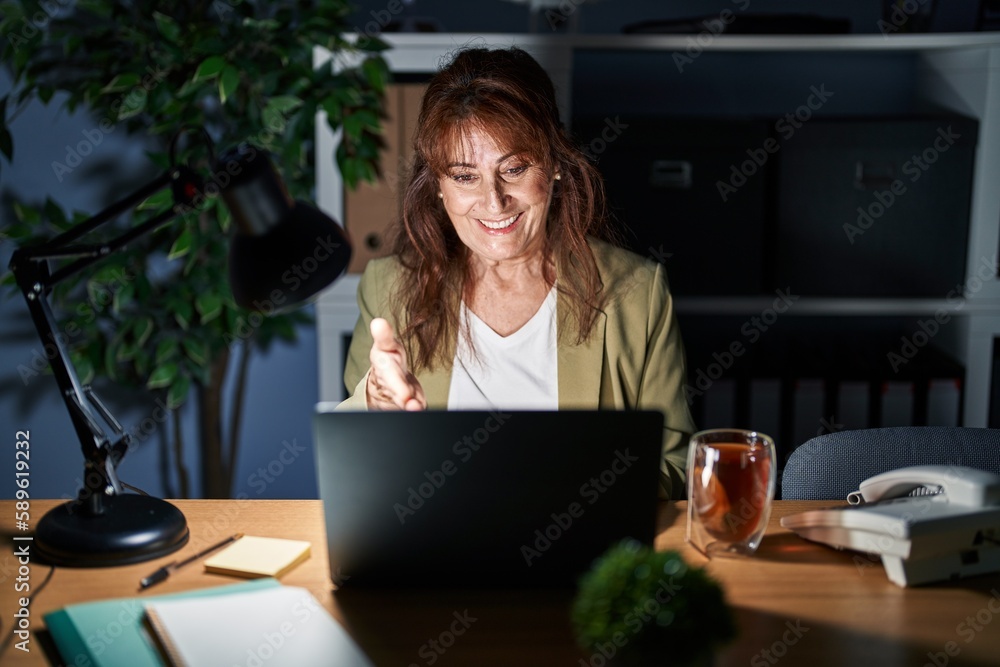 Middle age hispanic woman working using computer laptop at night smiling friendly offering handshake as greeting and welcoming. successful business. Middle age hispanic woman working using computer laptop at night smiling friendly offering handshake as greeting and welcoming. successful business.