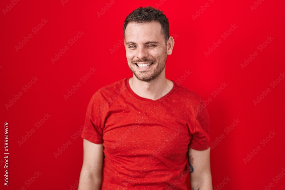 Obraz premium Young hispanic man standing over red background winking looking at the camera with sexy expression, cheerful and happy face.