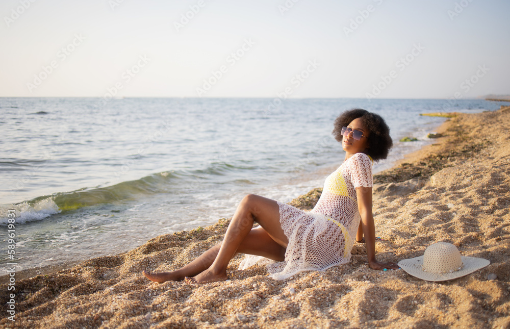Beautiful african-american woman relaxing on the beach with an afro hairstyle. Summer day, vacation, weekend, mood, yellow swimsuit, black leather, sunglasses, hat