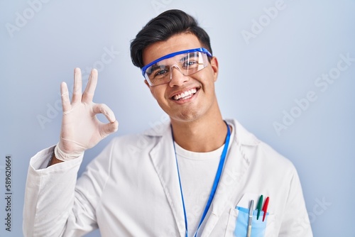 Hispanic man working as scientist smiling positive doing ok sign with hand and fingers. successful expression.