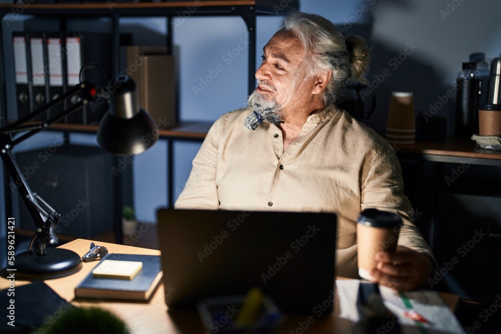 Middle age man with grey hair working at the office at night looking away to side with smile on face, natural expression. laughing confident. Middle age man with grey hair working at the office at night looking away to side with smile on face, natural expression. laughing confident.