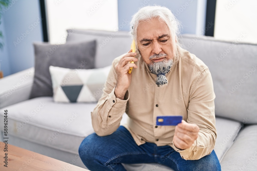 Middle age grey-haired man talking on smartphone holding credit card sitting on sofa at home Middle age grey-haired man talking on smartphone holding credit card sitting on sofa at home