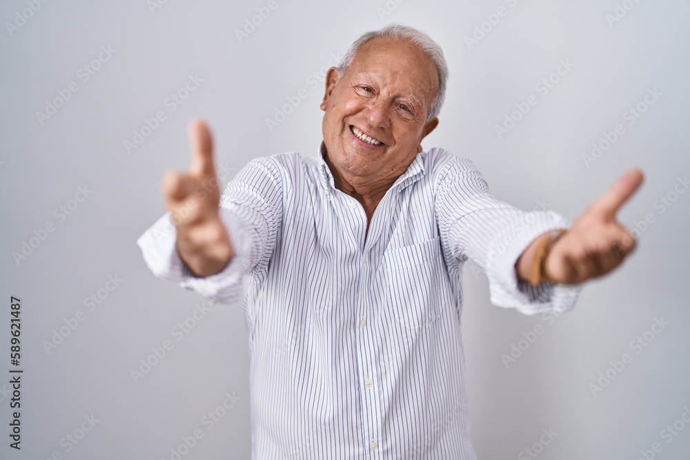 Senior man with grey hair standing over isolated background looking at the camera smiling with open arms for hug. cheerful expression embracing happiness. Senior man with grey hair standing over isolated background looking at the camera smiling with open arms for hug. cheerful expression embracing happiness.