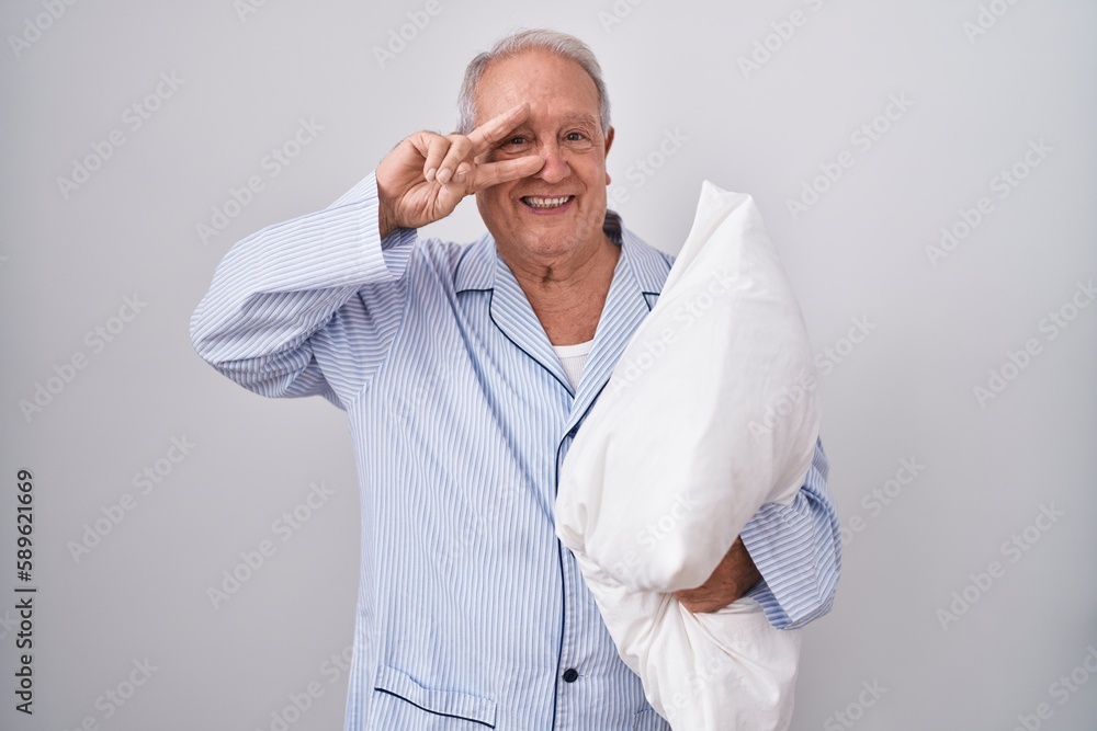 Senior man with grey hair wearing pijama hugging pillow doing peace symbol with fingers over face, smiling cheerful showing victory Senior man with grey hair wearing pijama hugging pillow doing peace symbol with fingers over face, smiling cheerful showing victory