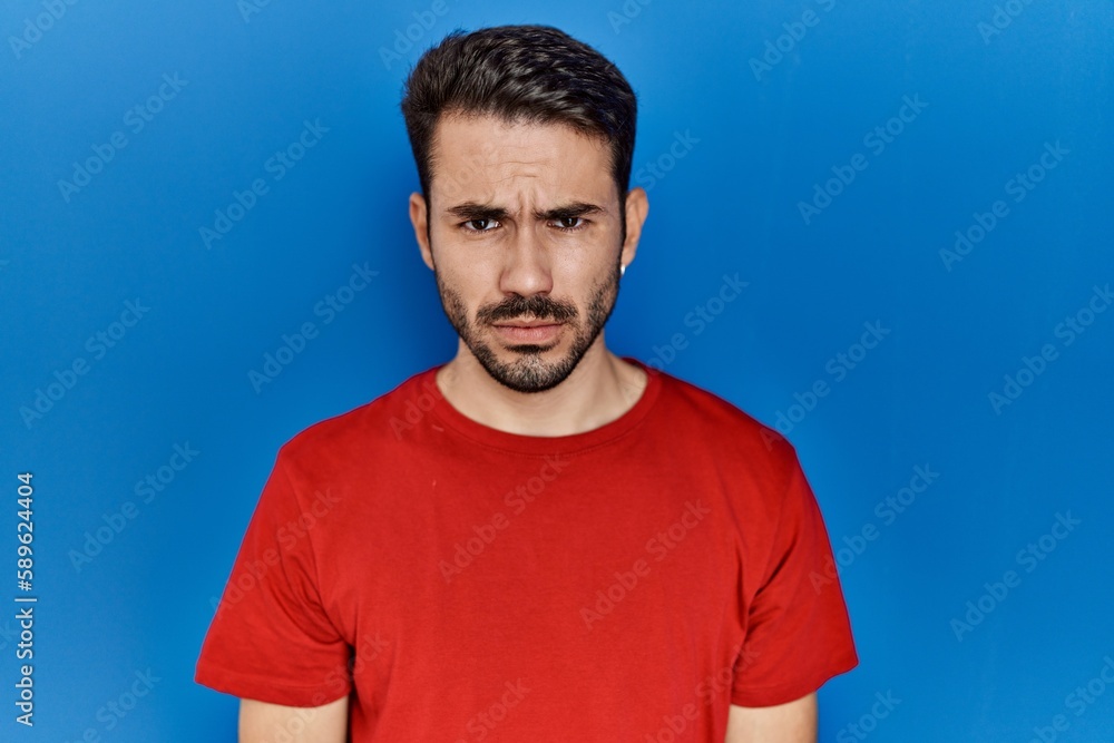 Young hispanic man with beard wearing red t shirt over blue background skeptic and nervous, frowning upset because of problem. negative person. Young hispanic man with beard wearing red t shirt over blue background skeptic and nervous, frowning upset because of problem. negative person.