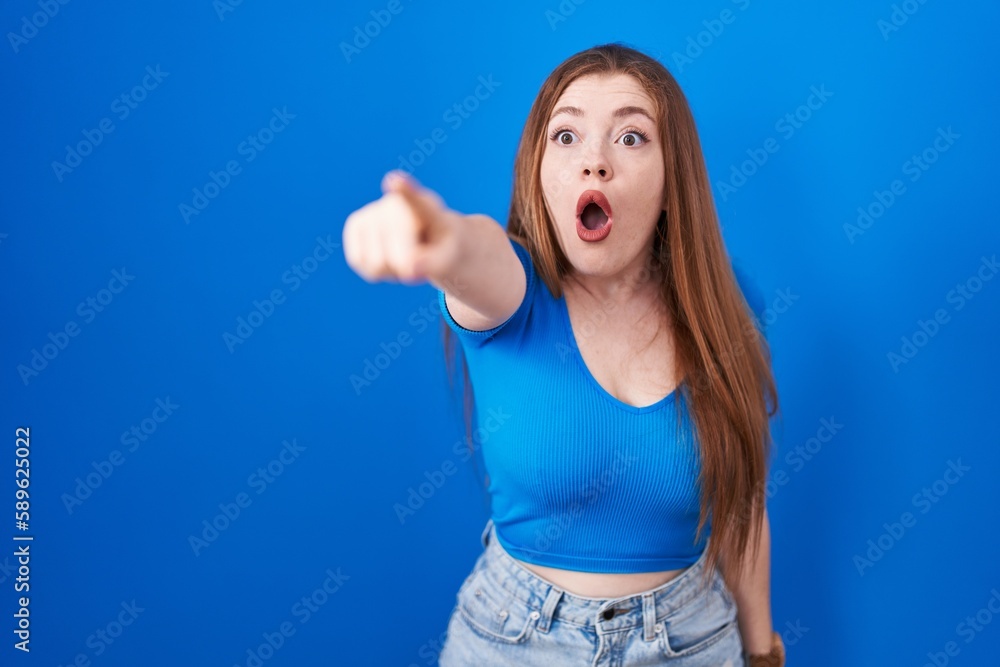 Redhead woman standing over blue background pointing with finger surprised ahead, open mouth amazed expression, something on the front