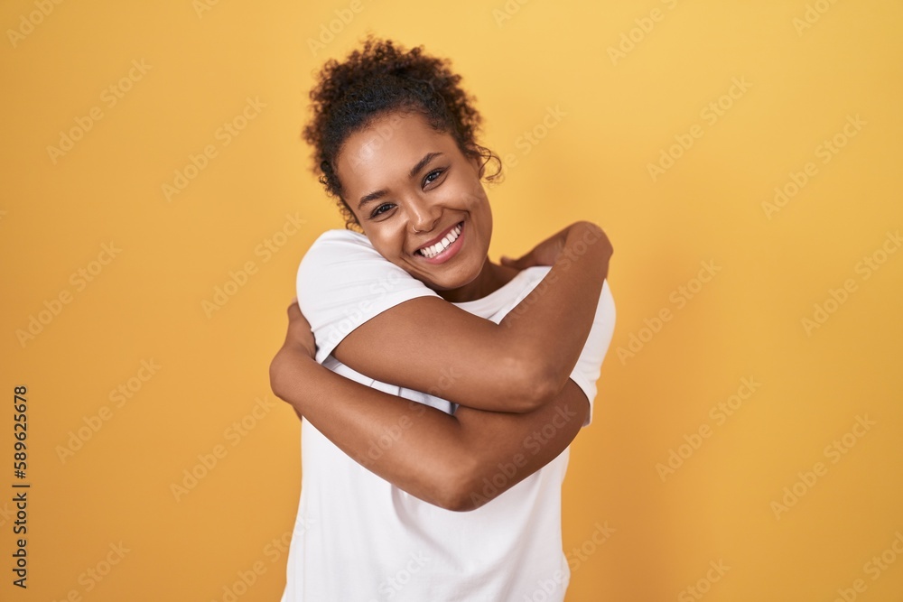 Young hispanic woman with curly hair standing over yellow background hugging oneself happy and positive, smiling confident. self love and self care Young hispanic woman with curly hair standing over yellow background hugging oneself happy and positive, smiling confident. self love and self care