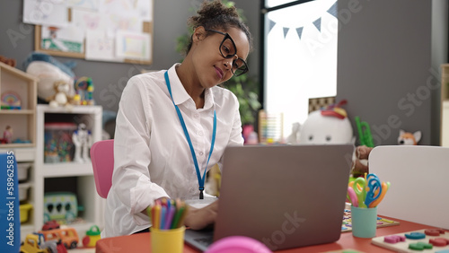 African american woman preschool teacher using laptop sitting on chair at kindergarten