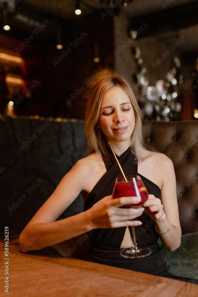 Portrait of young happy good-looking woman wearing black dress with long fair hair sitting at wooden table in restaurant, holding glass with red cocktail with straw. Celebration, party. Vertical.