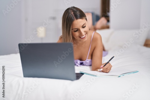 Young beautiful hispanic woman using laptop writing on notebook at bedroom