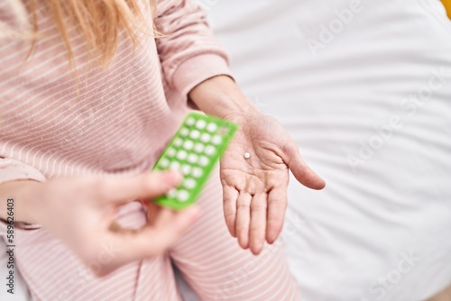 Young blonde woman holding birth control pills sitting on bed at bedroom