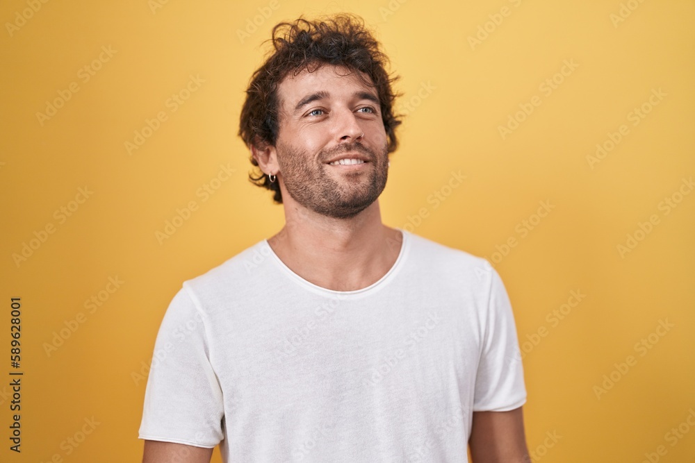 Young hispanic man smiling confident looking to the side over isolated yellow background Young hispanic man smiling confident looking to the side over isolated yellow background