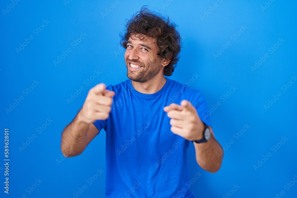Hispanic young man standing over blue background pointing fingers to camera with happy and funny face. good energy and vibes. Hispanic young man standing over blue background pointing fingers to camera with happy and funny face. good energy and vibes.