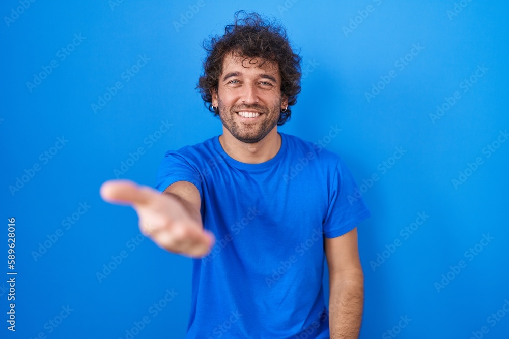 Naklejka premium Hispanic young man standing over blue background smiling cheerful offering palm hand giving assistance and acceptance.