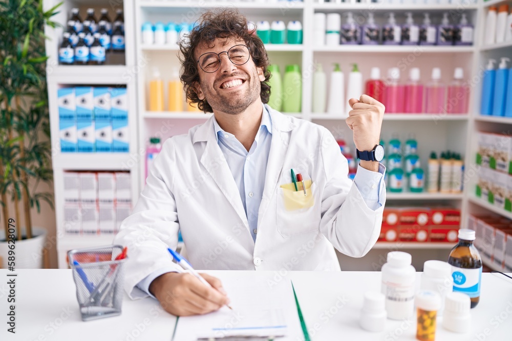 Hispanic young man working at pharmacy drugstore very happy and excited doing winner gesture with arms raised, smiling and screaming for success. celebration concept.