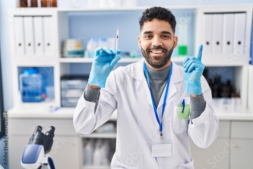 Hispanic man with beard working at scientist laboratory holding syringe smiling happy pointing with hand and finger to the side