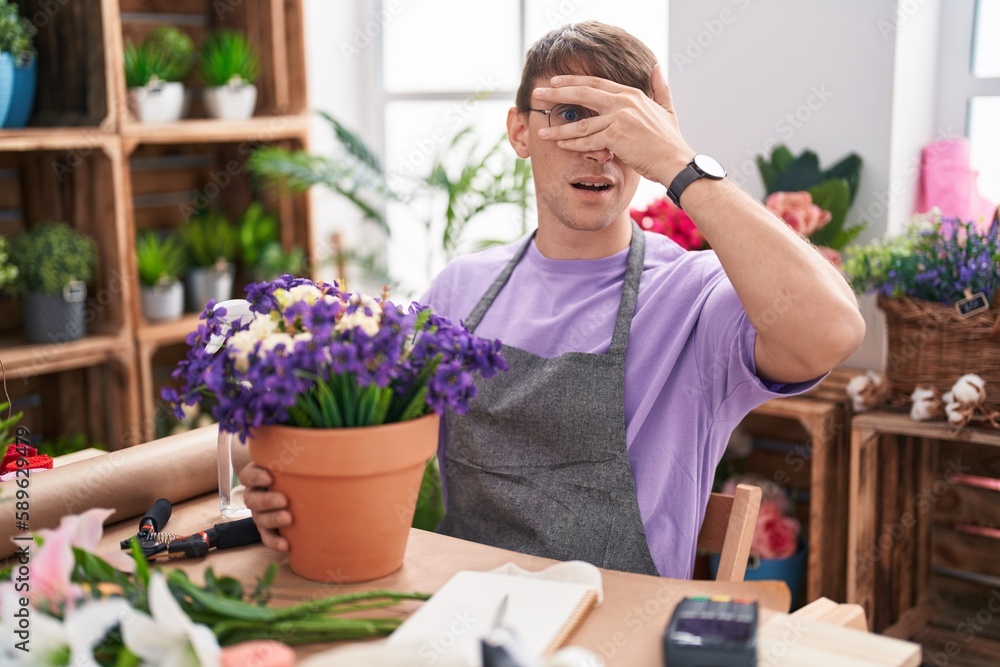 Caucasian blond man working at florist shop peeking in shock covering face and eyes with hand, looking through fingers with embarrassed expression.