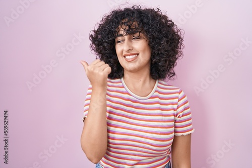 Young middle east woman standing over pink background smiling with happy face looking and pointing to the side with thumb up. © Krakenimages.com