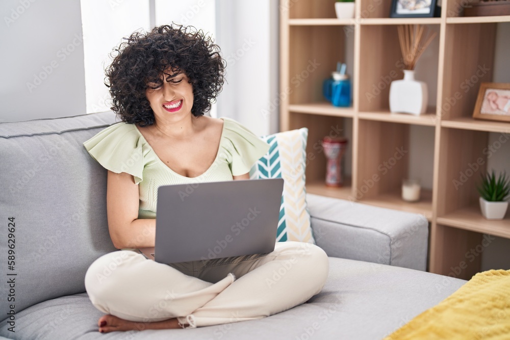 Young brunette woman with curly hair using laptop sitting on the sofa at home with hand on stomach because indigestion, painful illness feeling unwell. ache concept. Young brunette woman with curly hair using laptop sitting on the sofa at home with hand on stomach because indigestion, painful illness feeling unwell. ache concept.
