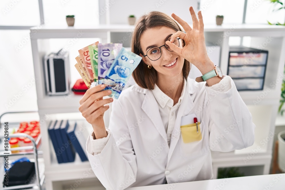 Young caucasian doctor woman holding canadian dollars banknotes smiling happy doing ok sign with hand on eye looking through fingers Young caucasian doctor woman holding canadian dollars banknotes smiling happy doing ok sign with hand on eye looking through fingers